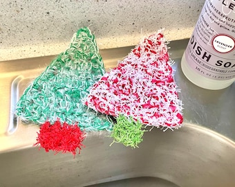 Puede incluir: Dos estropajos hechos a mano con forma de árbol de Navidad, en verde y rojo con detalles blancos, y dos estropajos más pequeños en rojo y verde. Una botella de jabón para platos con aroma a lavanda es visible en el fondo.