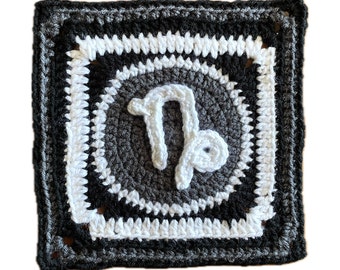 May include: A crocheted square with a black and white border and a gray center featuring the astrological symbol for Capricorn in white yarn.