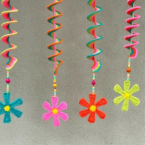 May include: Five colorful crocheted flower ornaments hanging from a ceiling. Each flower is a different color: teal, pink, orange, yellow, and green. The flowers are attached to long, colorful, crocheted strings.