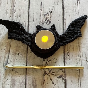 May include: Black crocheted bat with a small candle holder in the centre. The candle is lit and glowing yellow. The bat is on a white wooden background.