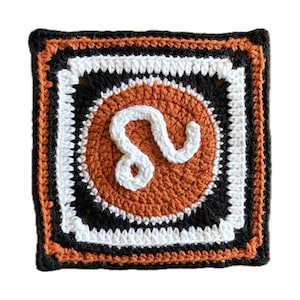 May include: Crocheted square featuring the astrological sign Leo in white yarn on a brown background. The square is framed with black and white yarn.