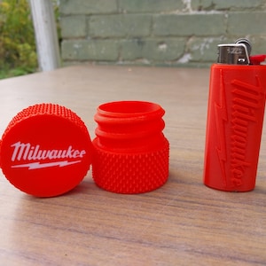 May include: A red lighter, a matching container, and a screw-on cap with the "Milwaukee" logo in white. The lighter and cap are cylindrical, and the container is a small, open-topped cylinder. The cap has a textured surface.
