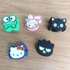 May include: Five colorful cartoon character charms. Includes a green frog, a pink bunny, a black cat, a white cat with a red bow, and a black bird. These charms are likely for shoes or accessories.