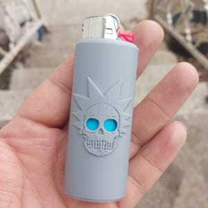 May include: Gray lighter with a skull design and blue eyes. The skull has a starburst pattern around it.