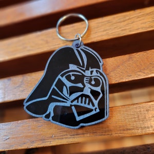 May include: A black and silver Darth Vader keychain made of wood. The keychain features a detailed silhouette of Darth Vader's helmet and face.