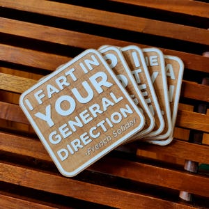 May include: Set of four square wooden coasters with rounded corners. Each coaster has the phrase "I FART IN YOUR GENERAL DIRECTION" in white text. The bottom of the coaster reads "-French Solider". The coasters are light brown.