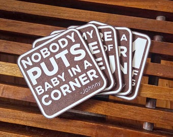 May include: A stack of brown square coasters with white text that reads "NOBODY PUTS BABY IN A CORNER -Johnny". The coasters are on a wooden surface.