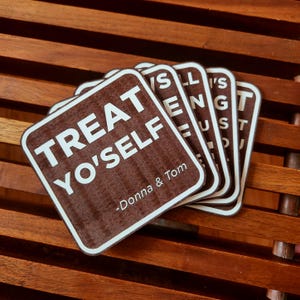 Puede incluir: Un montón de posavasos cuadrados marrones con texto blanco. El texto del primer posavasos dice "TREAT YO'SELF" y "-Donna & Tom". Otros posavasos muestran texto parcial. Esquinas redondeadas, borde blanco.