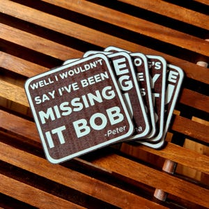 Peut inclure: Plusieurs dessous de verre carrés bruns avec du texte blanc. Le texte sur le premier dessous de verre se lit "WELL I WOULDN'T SAY I'VE BEEN MISSING IT BOB - Peter na". Les dessous de verre sont sur une surface en bois.