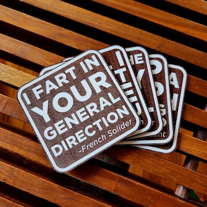 May include: Set of four square brown coasters with white text that reads "I FART IN YOUR GENERAL DIRECTION - French Solider". The coasters are stacked on a wooden surface.