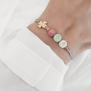 Könnte beinhalten: Ein personalisiertes Armband mit den Buchstaben "PIA" auf bunten Perlen. Das Armband hat einen goldfarbenen Kleeblatt-Anhänger und ist an einem grauen Band befestigt. Das Armband wird an einem Handgelenk vor weißem Hintergrund gezeigt.