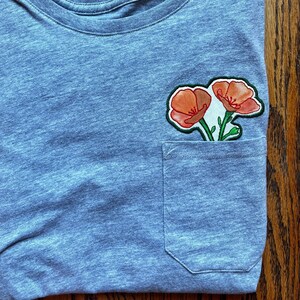 May include: A blue t-shirt with a pocket featuring an embroidered patch of two orange flowers.
