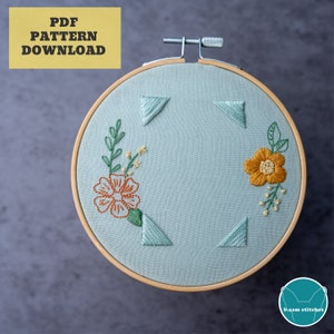 May include: A light blue embroidery hoop with a floral design. The design includes two orange flowers, green leaves, and small yellow flowers. The hoop is surrounded by a wooden frame.