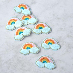 Peut inclure: Plusieurs écussons brodés en forme d'arc-en-ciel. Chaque patch présente un arc-en-ciel aux couleurs orange, jaune, rose et turquoise, sur des nuages bleus duveteux. Bordure dorée. Idéal pour personnaliser vêtements et accessoires.