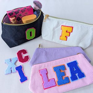 May include: A collection of corduroy cosmetic bags in various colors, each adorned with a large, embroidered letter. One black bag is open, revealing makeup items. Other bags are white, lavender, and pink, with letters in contrasting colors.