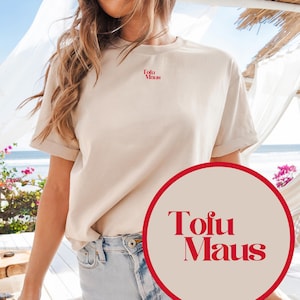 Könnte beinhalten: Beiges kurzärmeliges T-Shirt mit dem roten Text "Tofu Maus" auf der Brust und einer kleineren Version oben links. Das Shirt wird mit hellblauen Jeansshorts kombiniert. Das Bild wird im Freien mit Strandhintergrund aufgenommen.