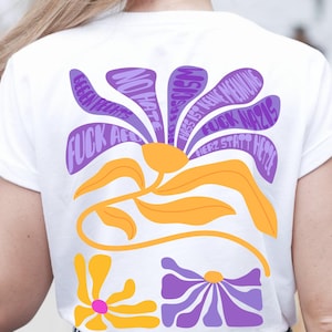 May include: White t-shirt featuring a floral design with purple and orange elements. The design includes text such as "NO HATE" and "FUCK NAZIS". The shirt has a crew neck and short sleeves.