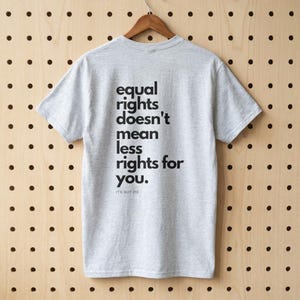 Può includere: Maglietta grigia con la scritta nera "equal rights doesn't mean less rights for you". La maglietta è appesa a una gruccia di legno su uno sfondo di legno con punti neri.