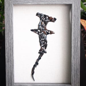 May include: A framed artwork featuring a hammerhead shark silhouette made from fossilized shark teeth. The teeth are arranged in a mosaic pattern, creating a unique and textured design. The frame is a simple gray wood.