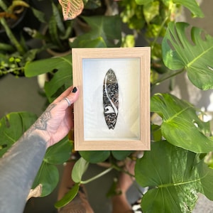 May include: A framed miniature surfboard with a black and white design. The surfboard is encased in a light brown wooden frame with a white background. The surfboard design features a wave and abstract patterns.