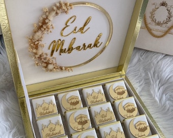 May include: A gold-colored box with a clear lid, filled with chocolates. Each chocolate is wrapped in white paper and topped with a gold-colored design and the words "Eid Mubarak". The box is decorated with a gold oval and dried flowers.