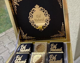 May include: A gold-trimmed box with a black interior, containing chocolates. The box lid features the words "Eid Mubarak" in gold script. The chocolates are decorated with gold accents and the same phrase.