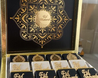 May include: A gold-trimmed box with a black interior featuring a gold floral design and the words "Eid Mubarak." Inside, individually wrapped chocolates are displayed, some with black and others with white wrappers, each adorned with gold "Eid Mubarak" accents.