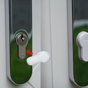 Puede incluir: Primer plano de una cerradura de puerta con un ojo de cerradura y una cubierta de llave blanca. La cerradura es plateada y está colocada sobre un fondo verde. El marco de la puerta es blanco. Una flecha roja apunta al ojo de la cerradura.