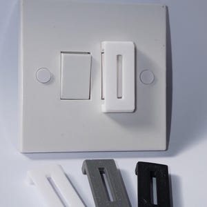 Könnte beinhalten: Eine weiße Lichtschalterabdeckung mit rechteckiger Öffnung, montiert auf einer weißen Wandplatte. Drei zusätzliche Abdeckungen in Weiß, Grau und Schwarz werden darunter angezeigt. Die Abdeckungen sollen eine versehentliche Schalterbetätigung verhindern.