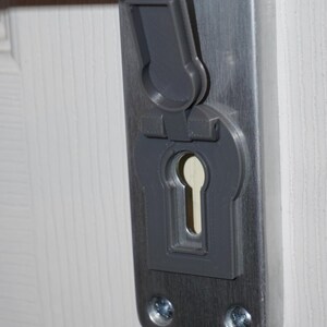 Könnte beinhalten: Eine graue Schlüssellochabdeckung an einer silbernen Türplatte. Die Abdeckung dient zum Schutz des Schlüssellochs und hat eine aufklappbare Klappe. Der Türgriff ist oben im Bild sichtbar. Die Türplatte ist an einer weißen Tür befestigt.
