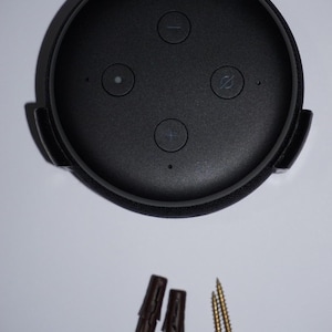 May include: A black smart speaker with control buttons and a circular design, mounted in a black holder. Includes two brown wall anchors and two gold screws for installation. The speaker has a matte finish.