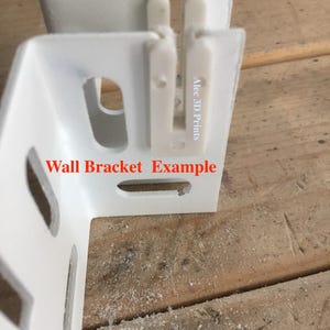 May include: A white, L-shaped wall bracket example with rectangular cutouts and a textured surface. The text "Wall Bracket Example" is printed in red. The bracket has a 3D printed appearance and is set against a wooden surface.
