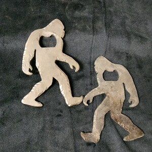 May include: Two metal bottle openers shaped like Bigfoot, one larger than the other. Both are silver in color and have a textured surface.