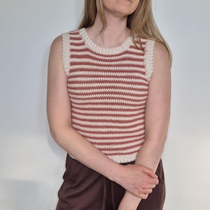 Op de afbeelding: Een mouwloze gehaakte top met horizontale strepen in terracotta en crème. De top heeft een ronde halslijn en een getailleerd silhouet. De haaksteek creëert een textuur. Het kledingstuk is handgemaakt.