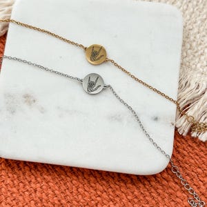 May include: Two delicate bracelets, one gold-toned and one silver-toned, each featuring a round charm with a hand gesture design. The bracelets rest on a white marble surface against a textured orange background.