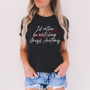 Peut inclure: T-shirt noir avec un texte blanc qui dit "I'd rather be watching Grey's Anatomy" avec un cœur rouge et un graphique de battement de cœur rouge.