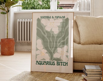 May include: A framed art print featuring a floral design with white flowers and green leaves on a light green background. The text "SOCIABLE & POPULAR" is at the top, and "AQUARIUS BITCH" is at the bottom in green. The print is displayed in a room with natural light.