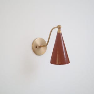 Op de afbeelding: Een wandlamp met een ronde messing muurplaat en een gebogen messing arm die een conische, glanzende terracotta-kleurige kap ondersteunt. Het armatuur heeft een gepolijste metalen afwerking op de bevestigingsonderdelen en een gladde, effen afwerking op de kegelvormige kap, die het licht naar beneden richt.