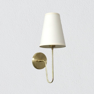 Op de afbeelding: Een messing wandlamp met een witte lampenkap van stof. De lamp heeft een gebogen arm en een ronde basis.