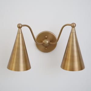 Op de afbeelding: Een messingkleurige wandlamp met twee conische kappen. De armatuur heeft een ronde achterplaat en gebogen armen die de kappen ondersteunen. De lamp is ontworpen voor binnenverlichting.