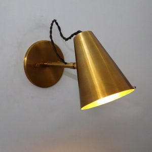 May include: A brass wall sconce with a cone-shaped shade. The sconce has a simple design and a warm, inviting glow.