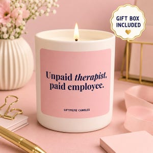 May include: A white cylindrical candle in a matte finish with a pink label featuring the dark blue text "Unpaid therapist, paid employee." and "GIFTMEME CANDLES" at the bottom. The candle has a lit wick and a gold-rimmed sticker stating "GIFT BOX INCLUDED".