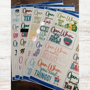 May include: Sheets of Avery labels with various phrases like "Open When You Need Things!" and "Open When You're Out of Funds!" in colorful fonts. Each label has a unique design, including illustrations of a shopping cart and money bags.