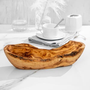 Può includere: Una ciotola di legno oblunga fatta a mano con una finitura marrone chiaro naturale. La ciotola presenta una forma organica unica e venature del legno visibili, perfetta per servire o esporre. La ciotola è lunga circa 35 cm.