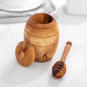 Può includere: Un vasetto di miele in legno con un coperchio e un mestolo per il miele. Il vasetto è fatto di legno marrone chiaro e ha una finitura in legno naturale. Il mestolo è anche fatto di legno e ha un design a nido d'ape.