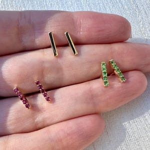 May include: A collection of gold-toned stud earrings. Two pairs are bar-shaped, and the other two pairs are rectangular with small, embedded gemstones. One pair has red gemstones, and the other has green gemstones.