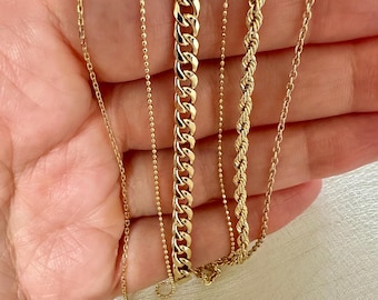 May include: A collection of gold necklaces in various styles. The necklaces include a delicate chain, a beaded chain, a curb chain, a rope chain, and a simple chain. The necklaces are displayed against a neutral background.