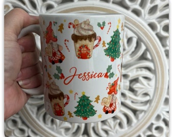 May include: White ceramic mug with a wraparound Christmas-themed design. The design includes gingerbread men, gnomes, Christmas trees, and hot chocolate mugs. The name "Jessica" is written in red script. The mug has a white handle.