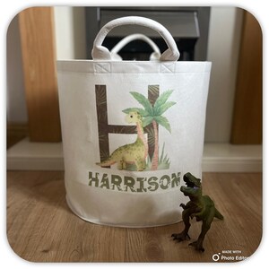May include: A white fabric storage basket with a dinosaur theme. The basket features a large letter "H" with a dinosaur and palm tree design. The name "HARRISON" is printed below the design. A green dinosaur toy is in front of the basket.