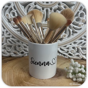 May include: A collection of makeup brushes in a white ceramic holder. The holder is inscribed with the name "Sienna" in black script, accompanied by a heart symbol. The brushes have silver handles and soft, light-colored bristles.